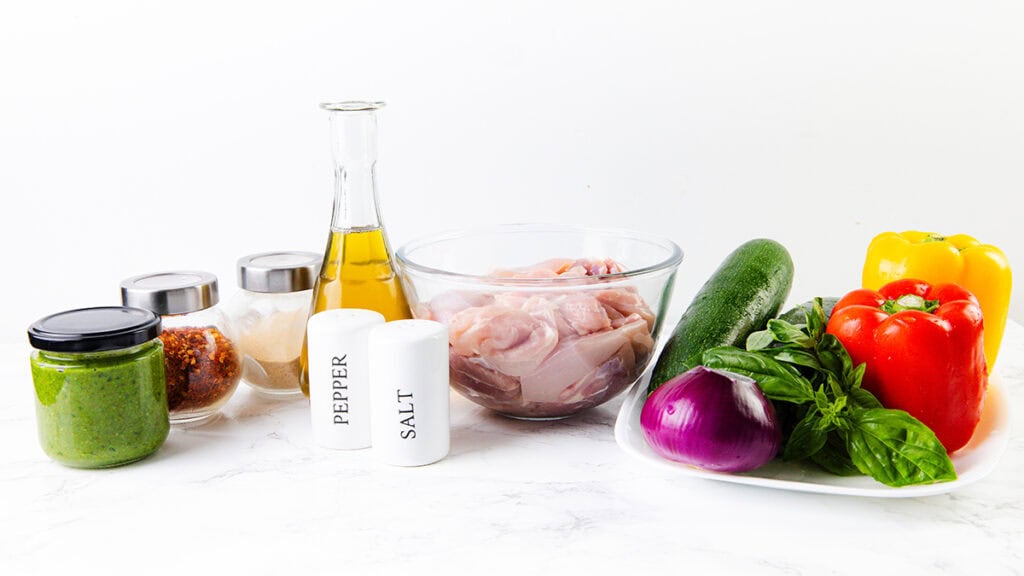 raw chicken pieces in a glass bowl, perfect for Pesto Chicken Sheet Pan meals—olive oil, salt, pepper, green sauce, seasonings in jars, zucchini, basil, red onion, and red and yellow bell peppers.