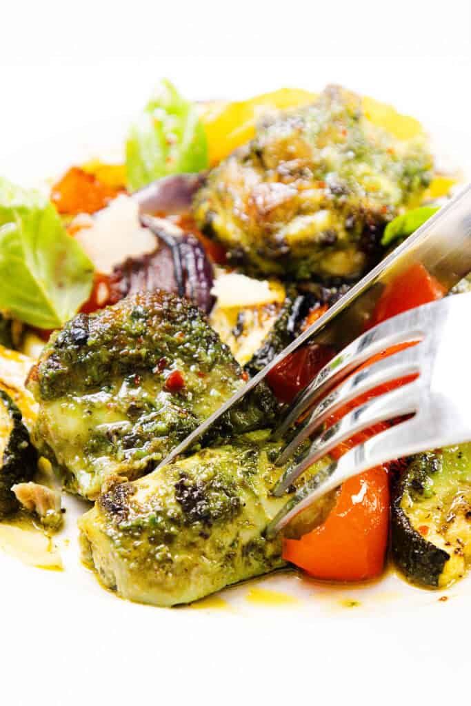A fork and knife cut into Pesto Chicken breast, surrounded by roasted sheet pan vegetables like zucchini, bell pepper, and leafy greens on a white plate.