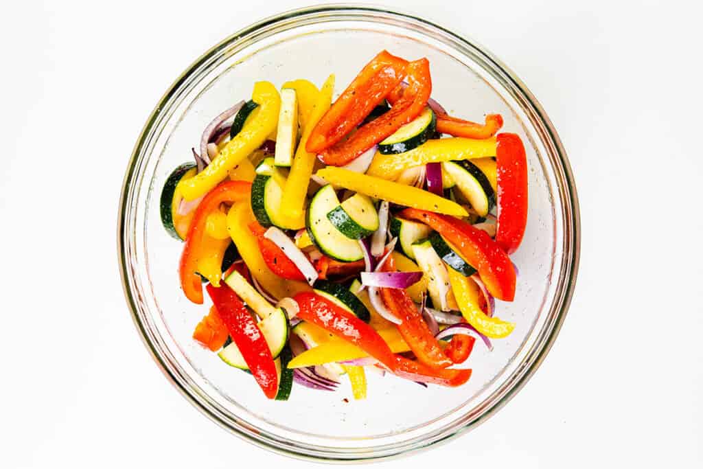 A glass bowl filled with sliced red and yellow bell peppers, zucchini, and red onion—perfect veggies to pair with sheet pan chicken—mixed together on a white background.