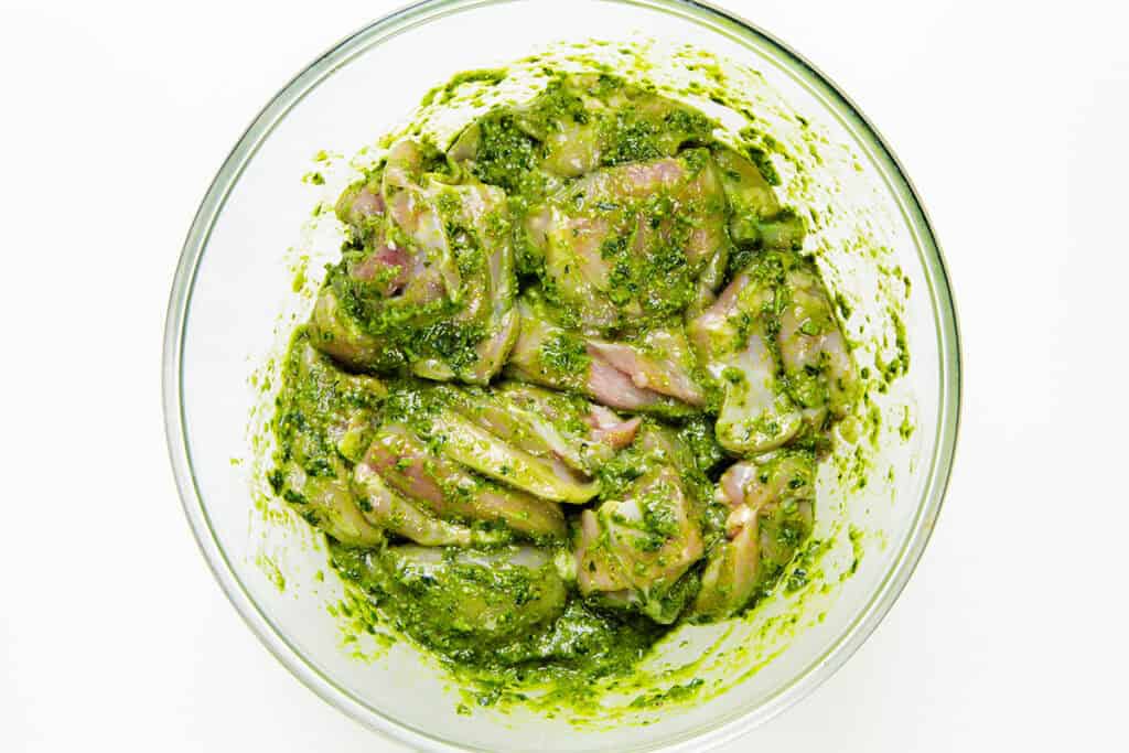 Raw chicken pieces coated in a vibrant green pesto marinade, perfect for making delicious Pesto Chicken, sit in a clear glass bowl on a white background.
