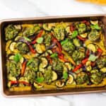 A sheet pan filled with pesto chicken meatballs and roasted vegetables, garnished with fresh basil leaves. Slices of bread and a jar of green sauce are nearby on a white marble surface.