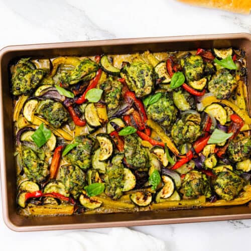 A sheet pan filled with pesto chicken meatballs and roasted vegetables, garnished with fresh basil leaves. Slices of bread and a jar of green sauce are nearby on a white marble surface.