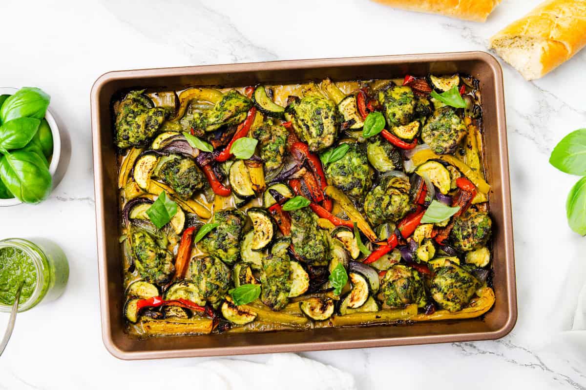 A sheet pan filled with pesto chicken meatballs and roasted vegetables, garnished with fresh basil leaves. Slices of bread and a jar of green sauce are nearby on a white marble surface.