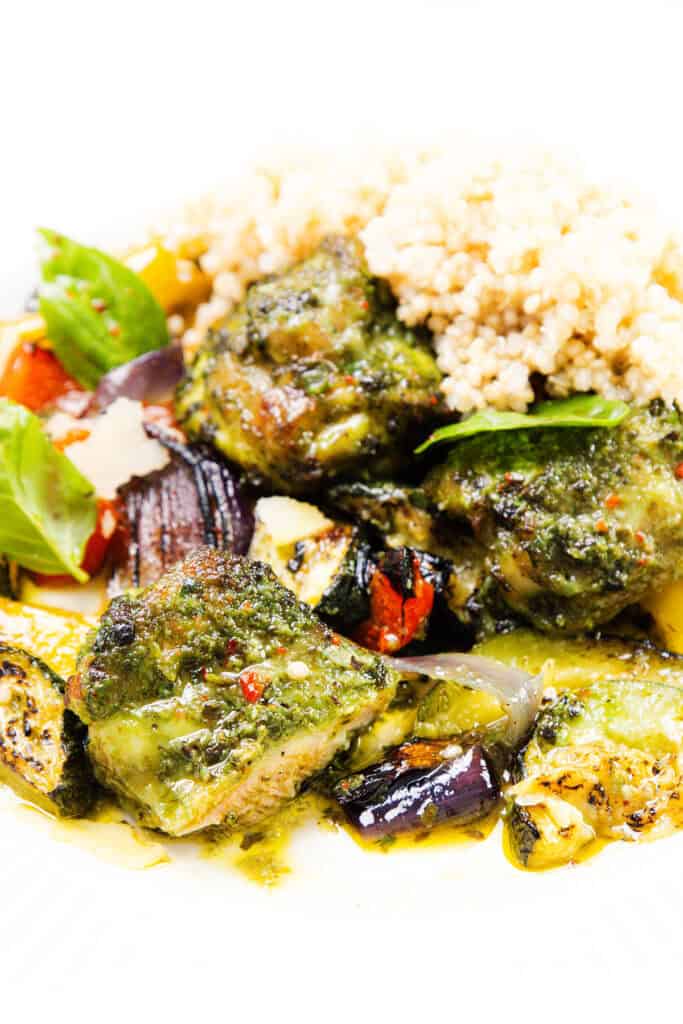 Colorful plate of pesto chicken with roasted tomatoes, zucchini, and red onion, garnished with fresh basil—a perfect sheet pan recipe—served alongside a portion of cooked quinoa in the background.