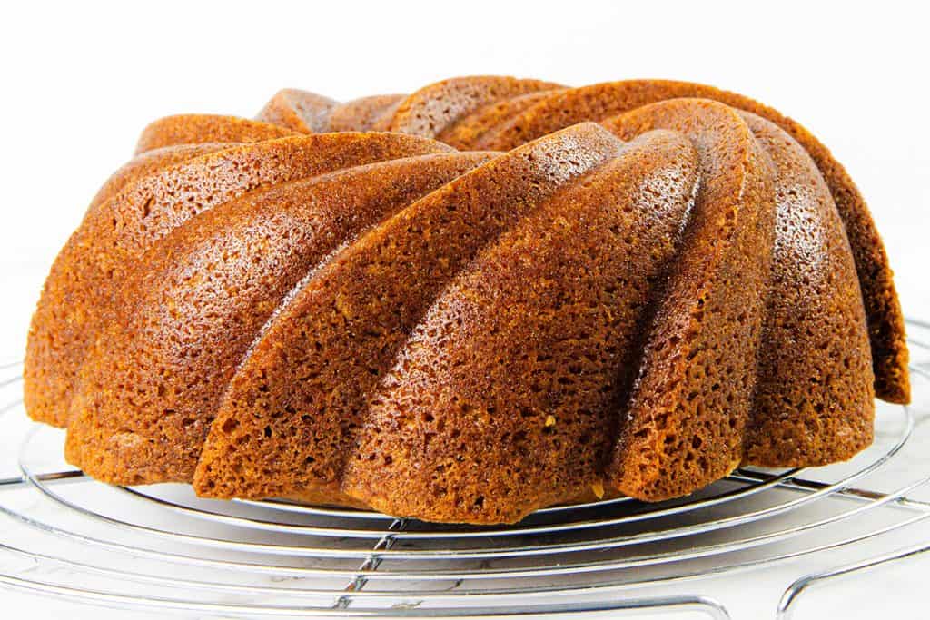A golden-brown Sock it to me cake with a swirled design sits on a round metal cooling rack against a white background, showcasing the classic bundt cake recipe.