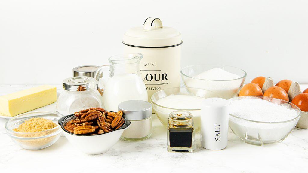 A variety of baking ingredients on a white surface, including eggs, flour, milk, butter, sugar, pecans, vanilla extract, salt, brown sugar, and cream—perfect essentials for your favorite cake recipe or a classic Sock it to me cake.