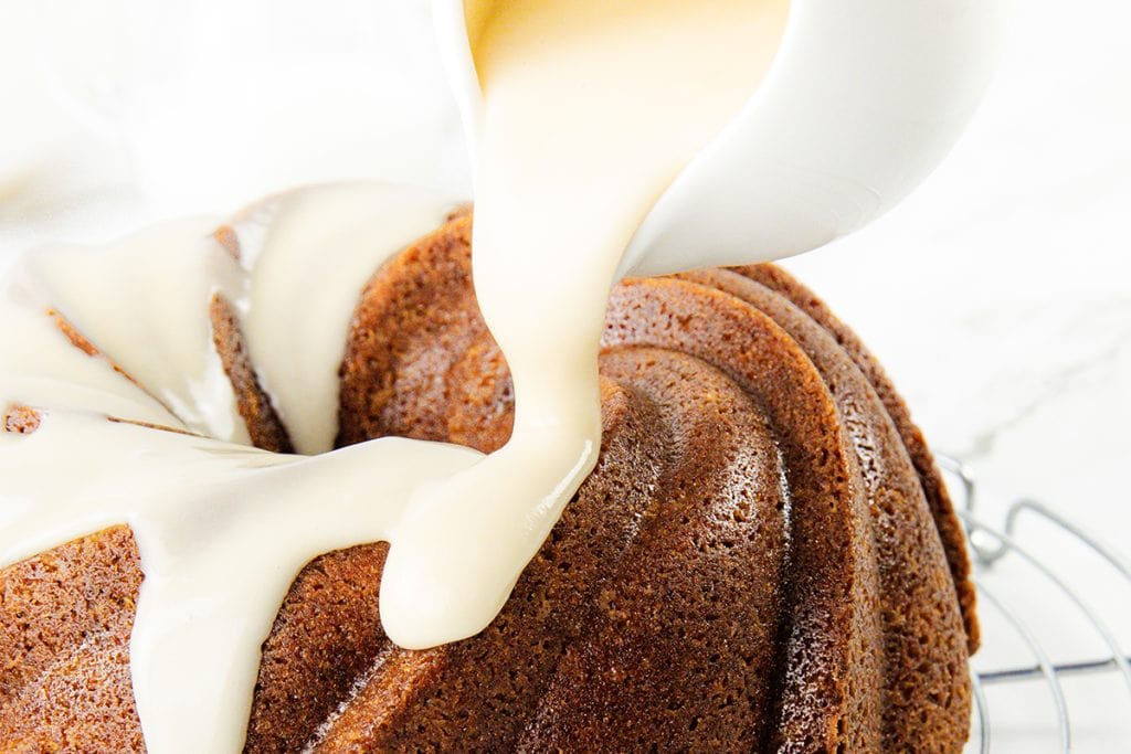 Close-up of thick, creamy white glaze being poured from a small white pitcher onto a Sock it to me cake resting on a wire cooling rack.