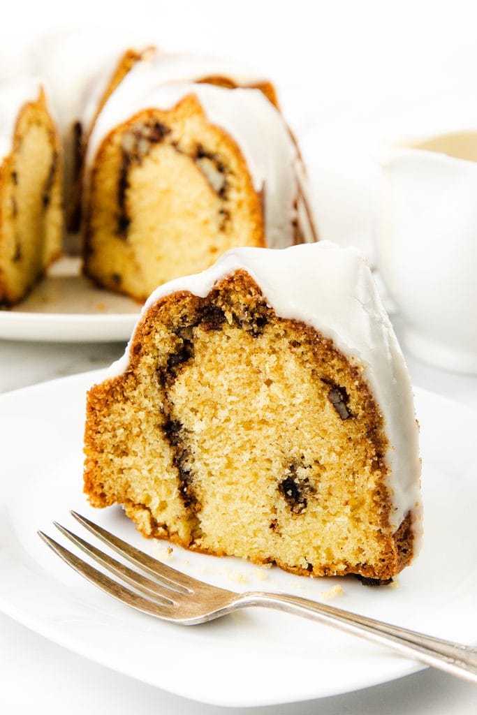 A slice of Sock it to me cake featuring a cinnamon swirl and white glaze sits on a white plate with a fork. The rest of the cake, partially sliced, can be seen in the background.