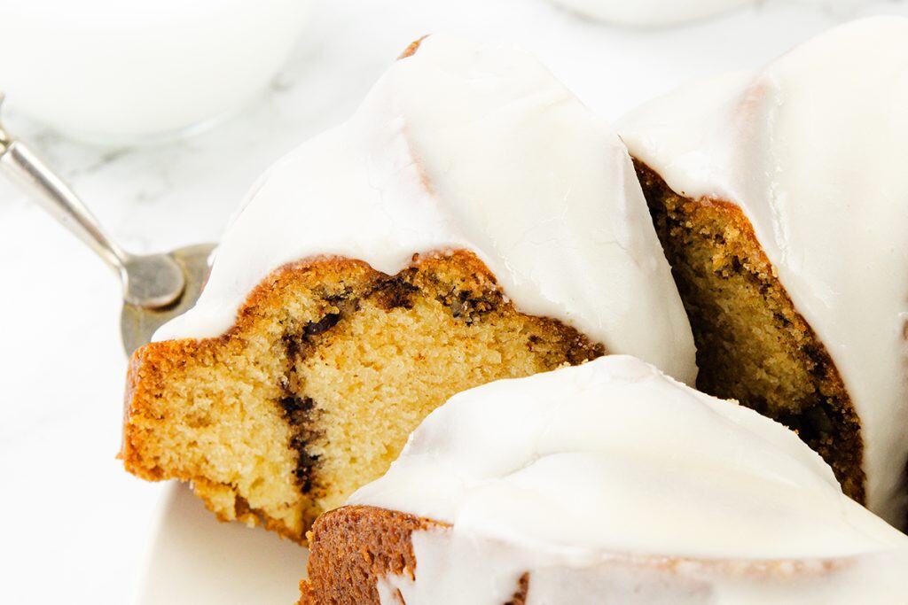 Close-up of slices of Sock it to me cake topped with a thick white glaze, showing a moist texture and a cinnamon ribbon inside. A fork and a glass of milk are partially visible in the background.