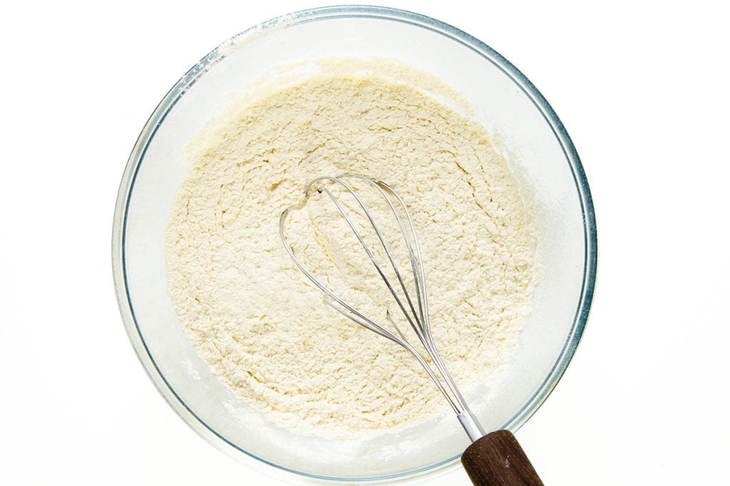 A glass bowl filled with flour mixture sits on a white surface. A metal whisk with a wooden handle rests inside the bowl, partially covered in the flour—perfect for starting your favorite Sock it to me cake recipe.