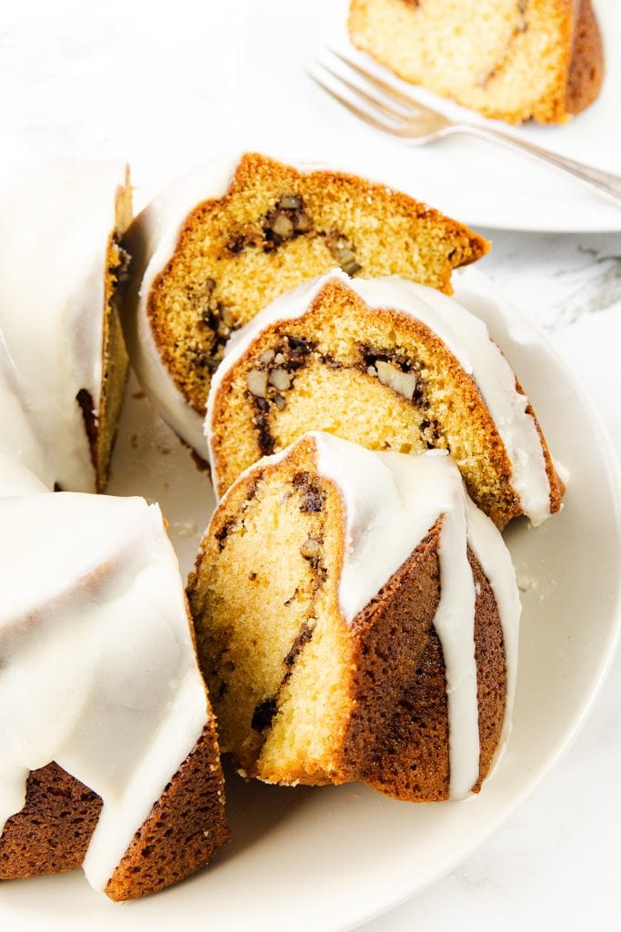 A sliced Sock it to me cake with white icing sits on a white plate. The bundt cake reveals a swirl of nuts and chocolate inside. In the background, a fork and another plate hold a piece of this delicious cake.