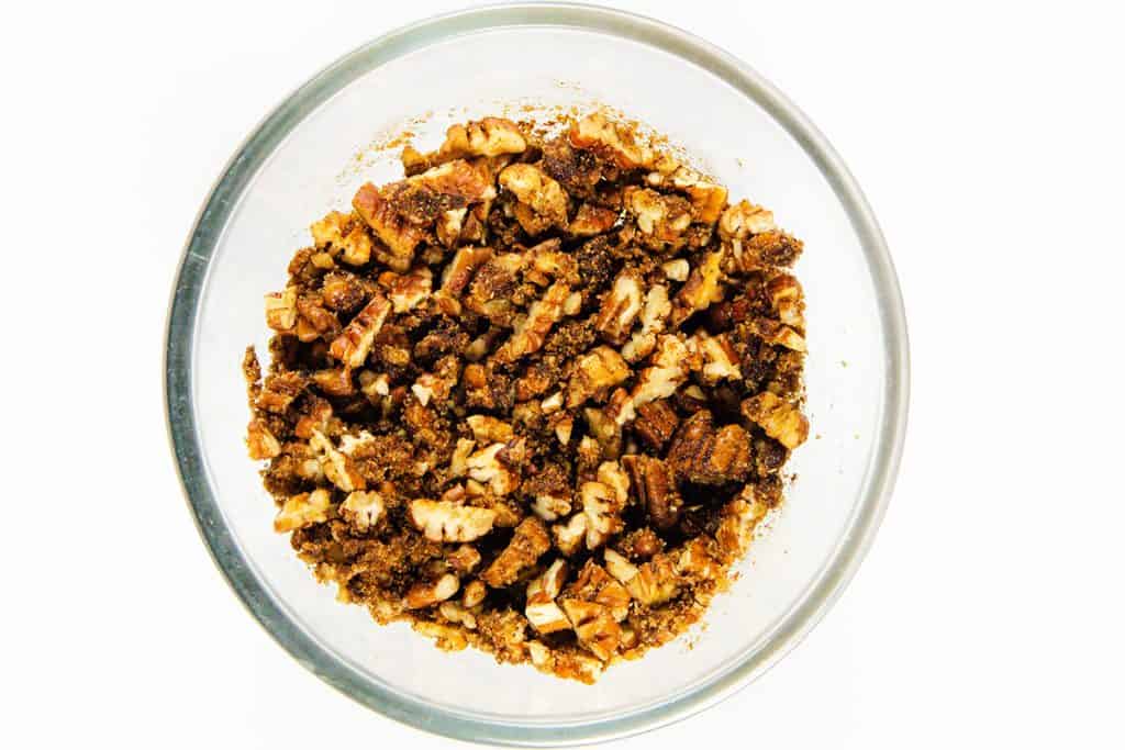 A glass bowl filled with chopped pecans mixed with brown sugar and spices, perfect for adding a crunchy layer to your favorite Sock it to me cake or cinnamon bundt cake, sits on a white background.