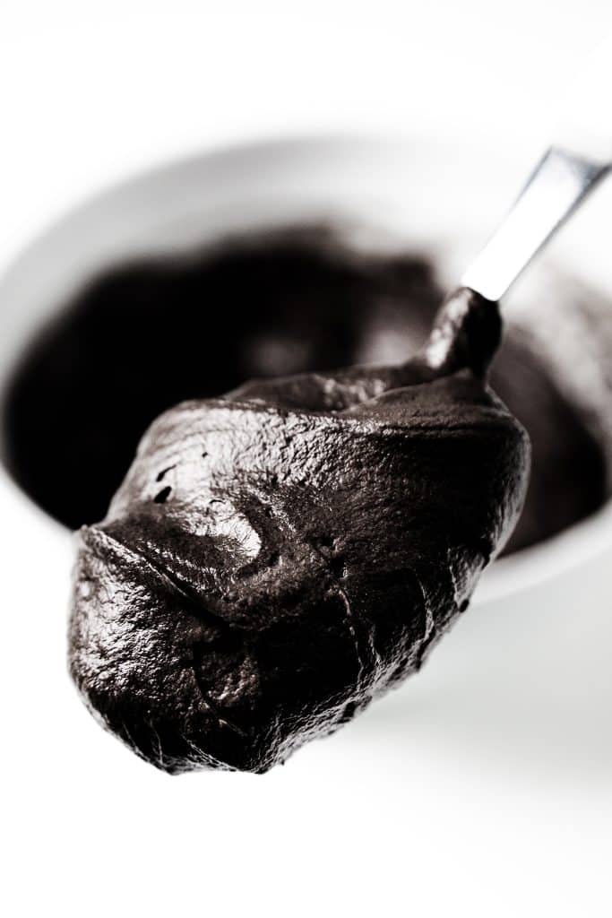 A close-up of a spoon holding thick, dark black chocolate frosting, with a bowl of more frosting blurred in the background.