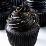 Three black cupcakes with glossy, swirled black frosting are shown. The cupcakes are in black wrappers and decorated with a light dusting of edible glitter and small gold accents, displayed on a white background.