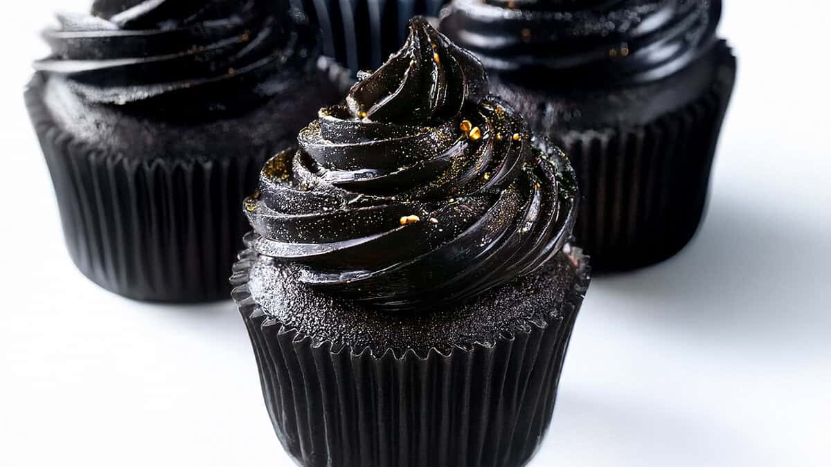 Three black cupcakes with glossy, swirled black frosting are shown. The cupcakes are in black wrappers and decorated with a light dusting of edible glitter and small gold accents, displayed on a white background.