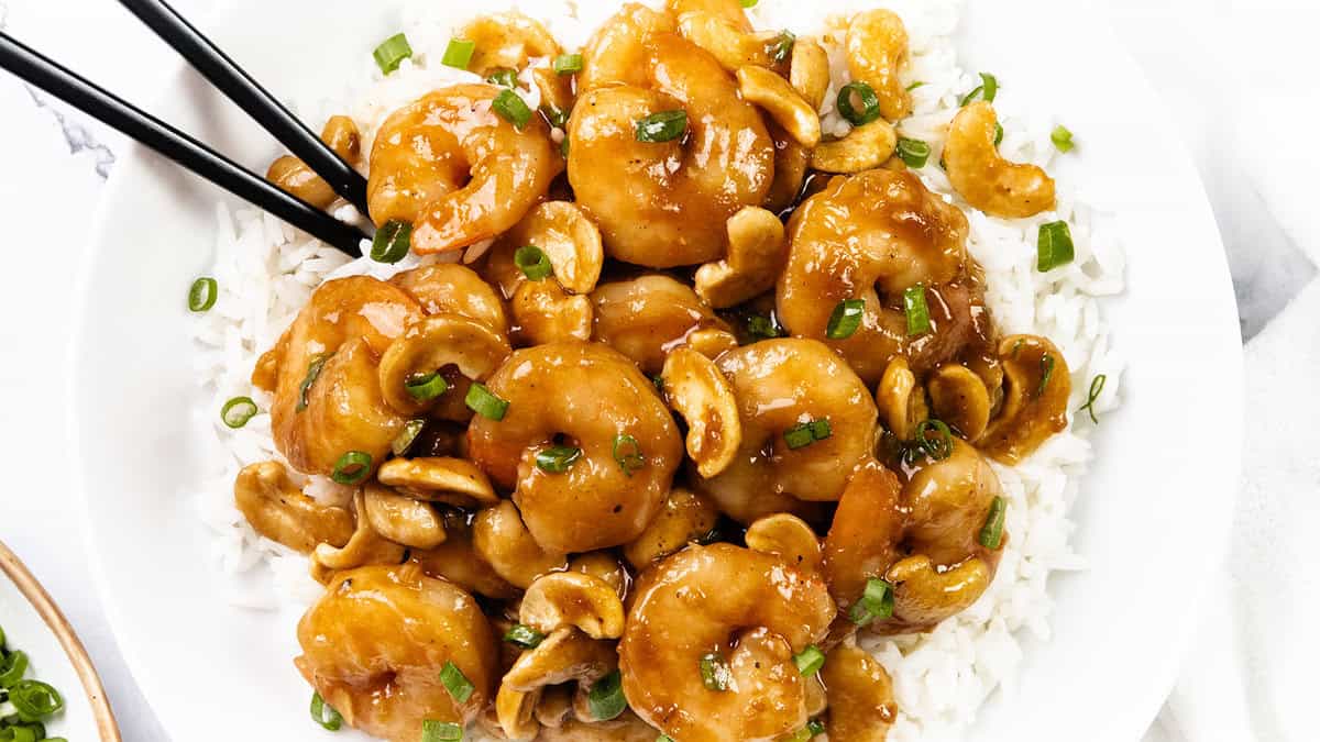 A white plate filled with white rice topped with shrimp, cashew nuts, and green onions in a brown sauce, with black chopsticks picking up some shrimp.
