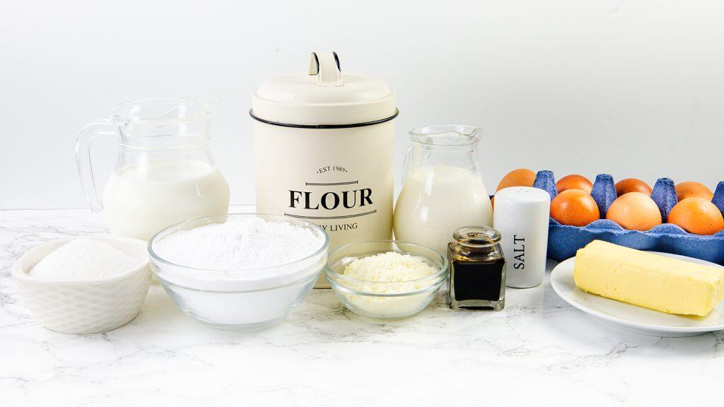 Various baking ingredients on a white surface, including milk, flour, sugar, grated cheese, vanilla extract, salt, eggs in a carton, and a stick of butter.