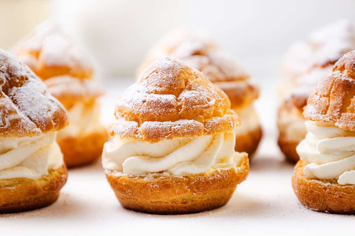 Close-up of several cream puffs topped with powdered sugar, filled with swirls of whipped cream, arranged on a white surface with a blurred background.