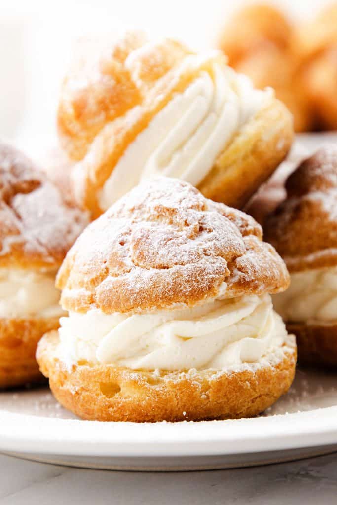 Close-up of cream puffs filled with whipped cream and dusted with powdered sugar, arranged on a white plate.