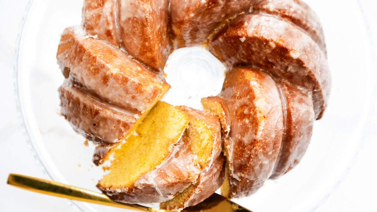 A glazed bundt cake with a golden interior sits on a glass cake stand. A gold knife is partially inserted, lifting two thick slices from the cake.