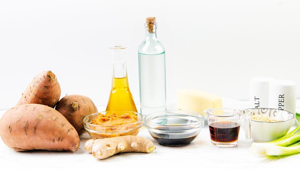 A selection of ingredients for Miso-Glazed Sweet Potatoes on a white surface, including sweet potatoes, fresh ginger, miso paste, soy sauce, butter, oil, measuring cups with liquids, rolled oats, green onions, and salt and pepper shakers.