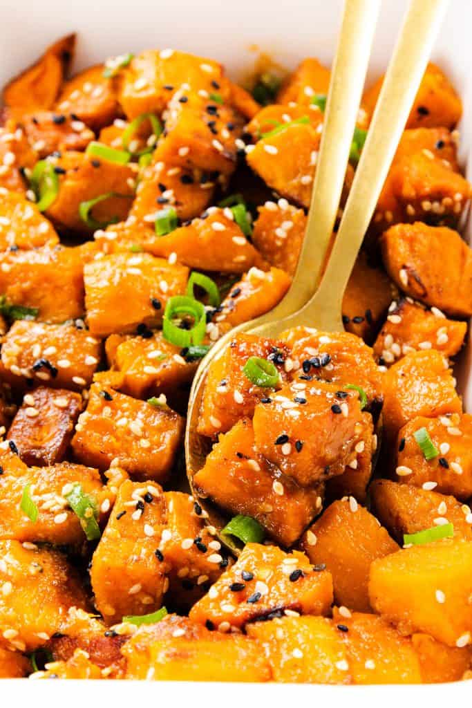 Close-up of miso-glazed sweet potatoes cubes garnished with chopped green onions, black and white sesame seeds, and served with two gold-colored utensils.
