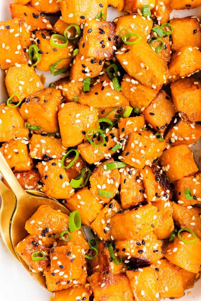 Close-up of Miso-Glazed Sweet Potatoes, roasted into cubes and topped with black and white sesame seeds and sliced green onions, served with a gold-colored spoon.