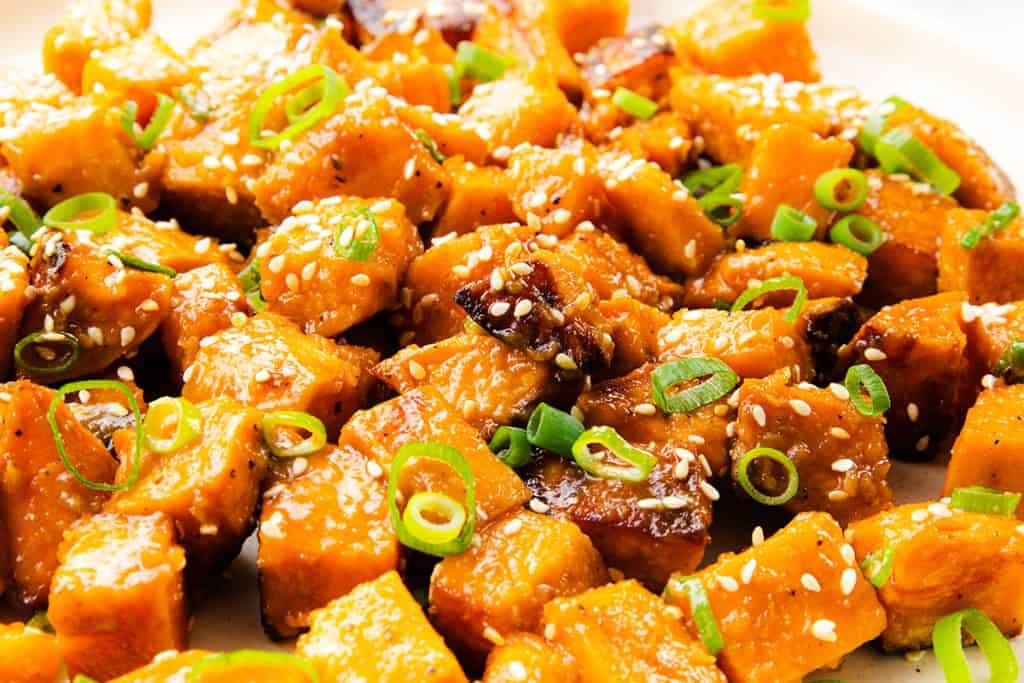 Close-up of Miso-Glazed Sweet Potatoes garnished with sliced green onions and sprinkled with sesame seeds on a white plate.