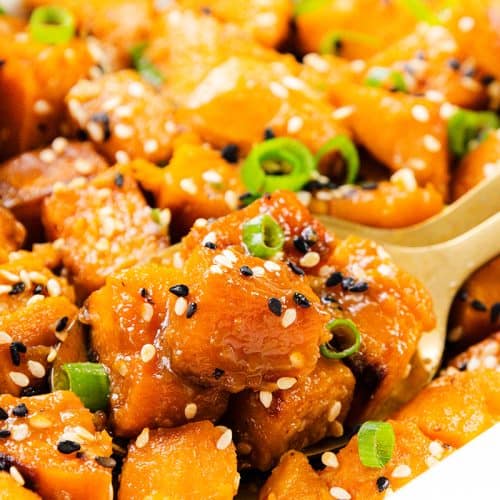 Close-up of Miso-Glazed Sweet Potatoes, roasted and garnished with black and white sesame seeds and chopped green onions, served with a gold spoon.