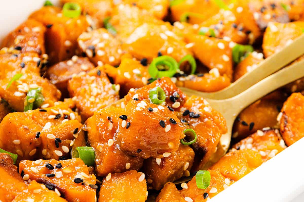 Close-up of Miso-Glazed Sweet Potatoes, roasted and garnished with black and white sesame seeds and chopped green onions, served with a gold spoon.
