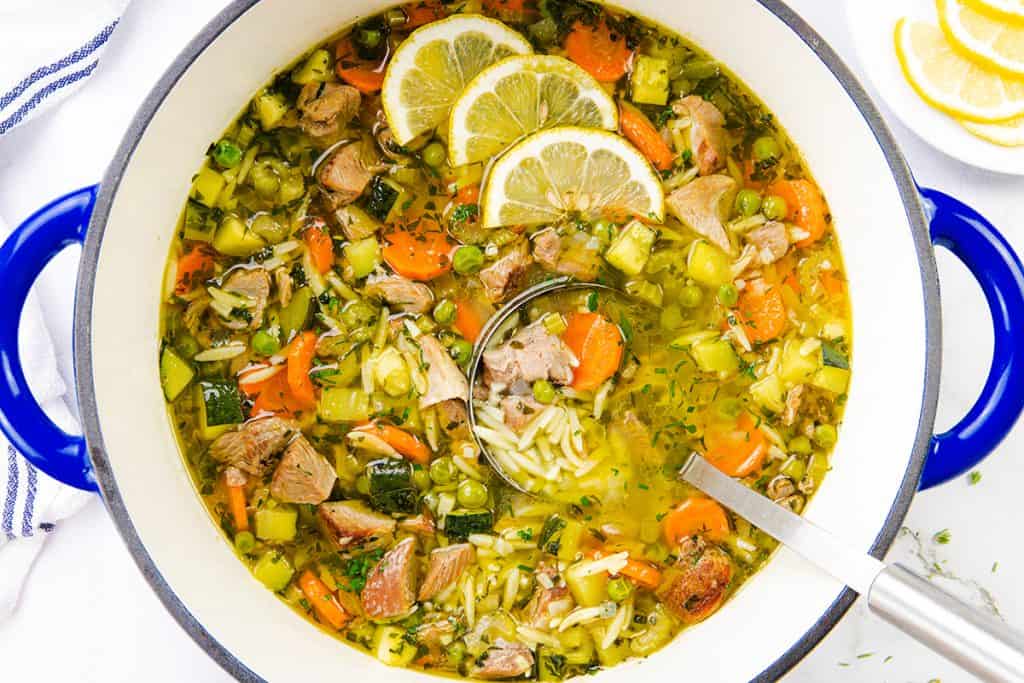 A vibrant one-pot orzo soup featuring chunks of leftover turkey, carrots, peas, and zucchini, garnished with lemon slices. A ladle rests inside the pot, with extra lemon slices on a plate nearby.
