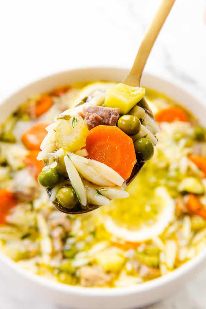 A close-up of a spoonful of Orzo Soup featuring peas, carrots, zucchini, and chunks of leftover turkey, held above a bowl filled with the same colorful one-pot meal.