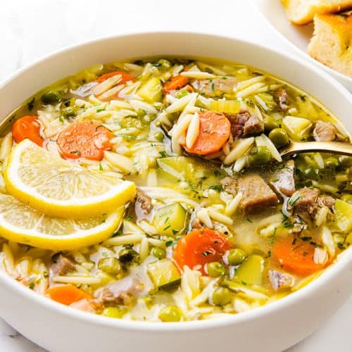 A bowl of one-pot orzo soup with carrots, celery, peas, leftover turkey chunks, and lemon slices, with a spoon inside. In the background, a plate of bread is partially visible.