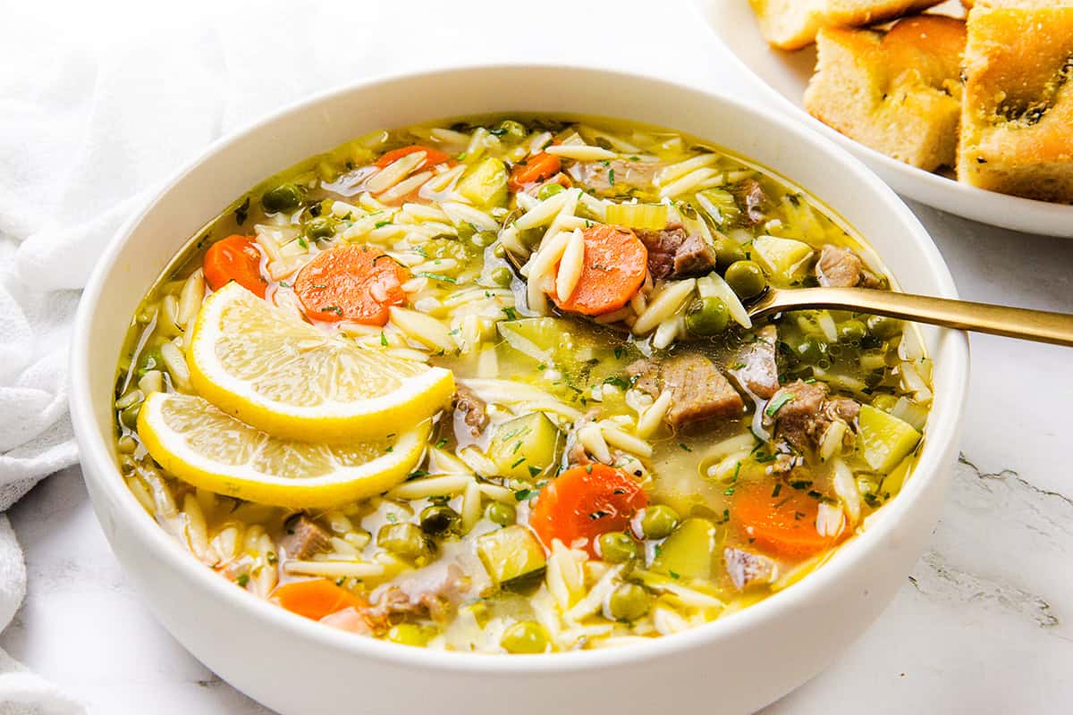 A bowl of one-pot orzo soup with carrots, celery, peas, leftover turkey chunks, and lemon slices, with a spoon inside. In the background, a plate of bread is partially visible.