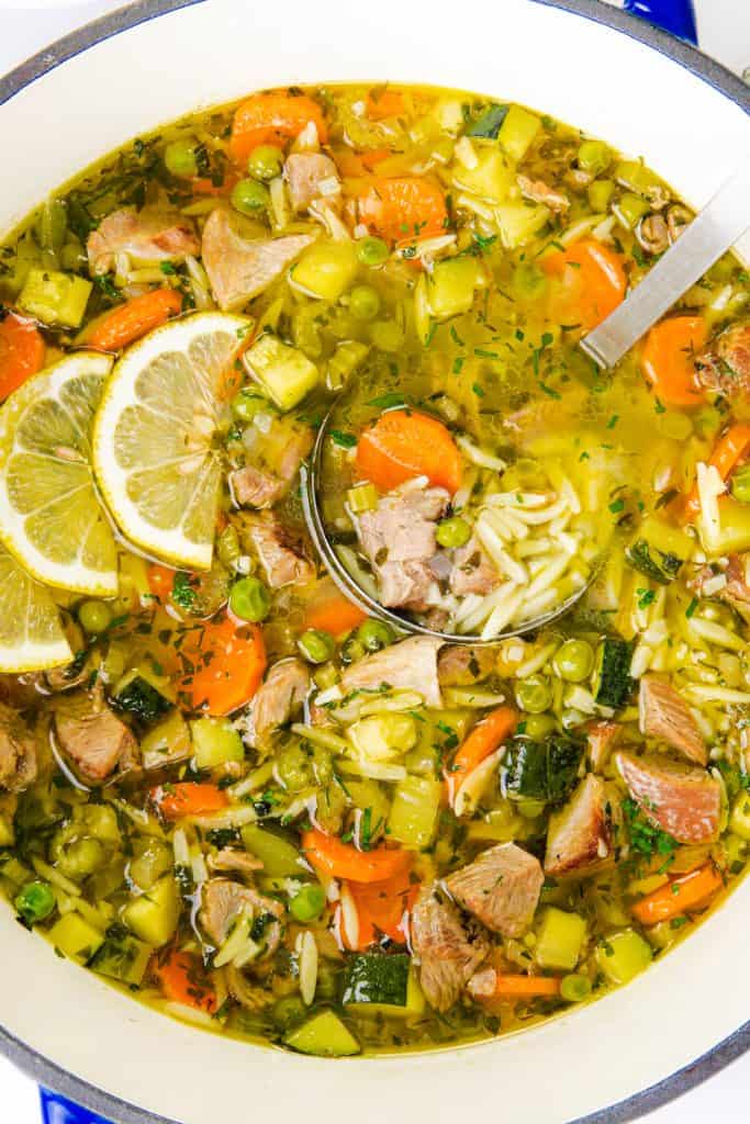 A one-pot orzo soup filled with leftover turkey, carrots, peas, zucchini, herbs, and slices of lemon. A ladle rests in the soup, revealing hearty chunks of meat and vegetables.