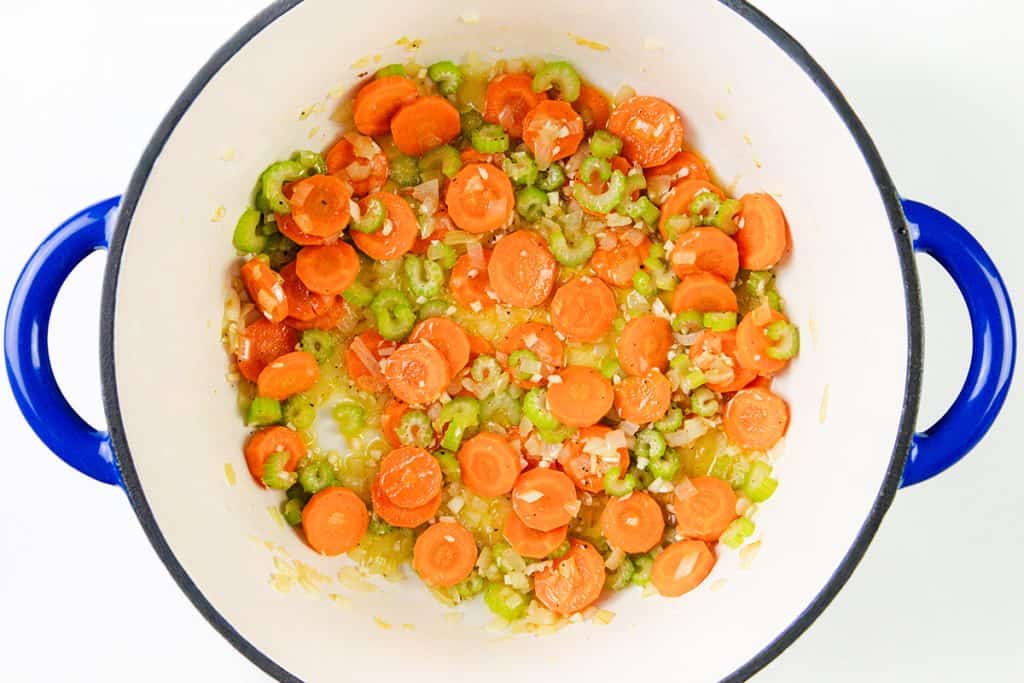 Sliced carrots, celery, and chopped onions sautéing in a white pot with blue handles—these veggies are lightly cooked and mixed together, perfect for the base of a hearty one-pot orzo soup or for using up leftover turkey.