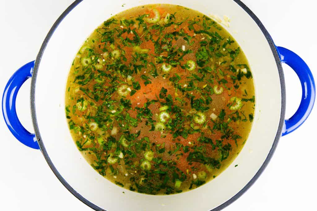 A blue-handled pot filled with a clear orzo soup, featuring leftover turkey, chopped green herbs, sliced okra, and chunks of tomato, viewed from above on a white background.