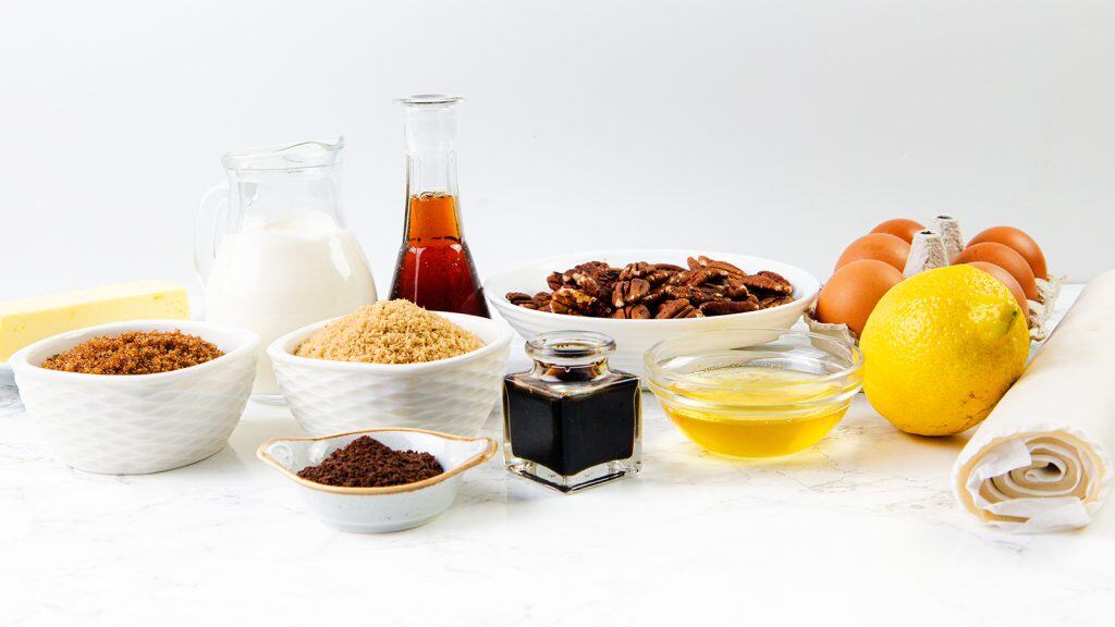 Assorted baking ingredients on a white surface, including eggs, milk, butter, brown sugar, vanilla, pecans, honey, a lemon, and a roll of pastry dough.