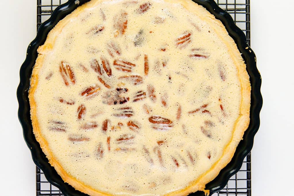 A pecan pie with a creamy topping and visible pecan halves sits in a round, fluted black pie dish on a metal cooling rack against a white background.