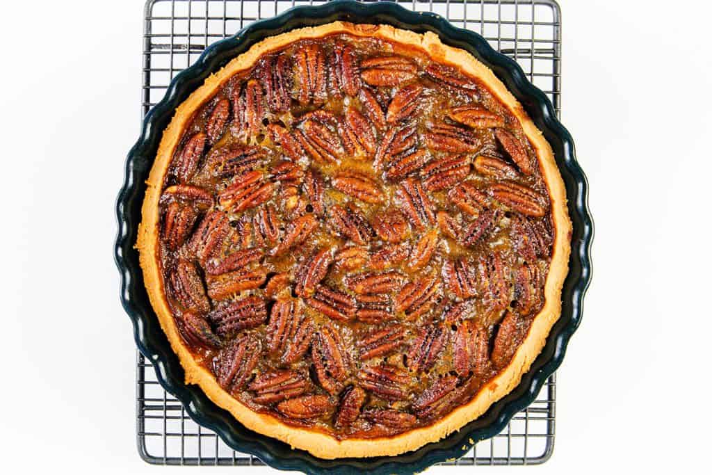 A freshly baked pecan pie in a round black pie dish sits on a cooling rack, viewed from above. The golden crust encircles a filling topped with whole glazed pecans.