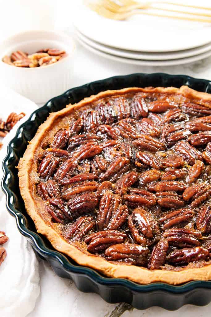 A close-up of a pecan pie in a black pie dish, topped with glossy pecans. In the background, a small white bowl with extra pecans and stacked white plates with gold forks can be seen.