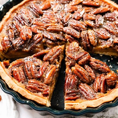 A close-up of a pecan pie with a golden crust in a black pie dish, with one slice slightly pulled out. Whole pecans cover the top, and a few pecans are scattered nearby on a white cloth.