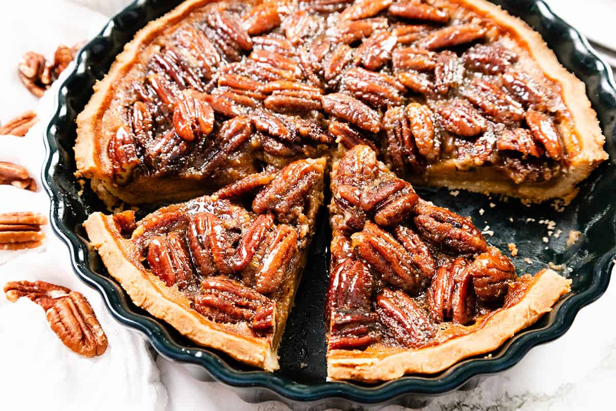 A close-up of a pecan pie with a golden crust in a black pie dish, with one slice slightly pulled out. Whole pecans cover the top, and a few pecans are scattered nearby on a white cloth.