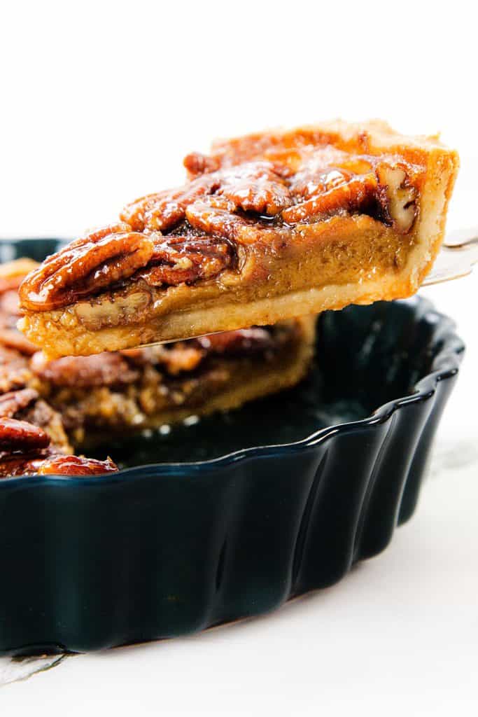 A close-up of a slice of pecan pie being lifted from a black pie dish, showing a thick, gooey filling topped with whole pecans and a flaky golden crust.
