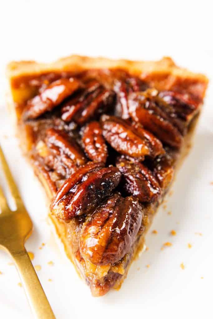 A close-up of a slice of pecan pie on a white plate, showing glossy, caramelized pecans on top of a golden crust, with a gold fork beside it.