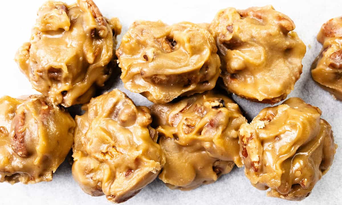 Seven pecan pralines with a creamy, caramel-colored coating are grouped together on a white surface. These classic 1980s desserts have a rough, chunky texture with visible pecan pieces throughout.
