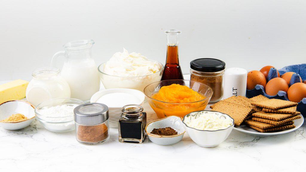 A variety of baking ingredients for Pumpkin Spice Cheesecake, including eggs, graham crackers, pumpkin puree, vanilla extract, sugar, spices, butter, cream cheese, and milk, arranged in bowls and jars on a white surface.