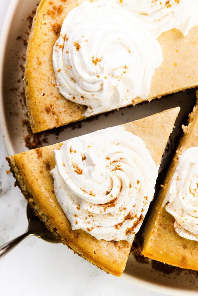 A close-up of a Pumpkin Spice Cheesecake topped with swirls of whipped cream and a sprinkle of cinnamon. One slice is being lifted from the whole cake on a plate.