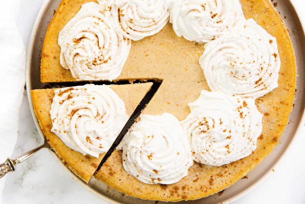 A Pumpkin Spice Cheesecake pie with six swirls of whipped cream and a sprinkle of cinnamon on top. One slice is being lifted out with a pie server, revealing the creamy, spiced texture inside.