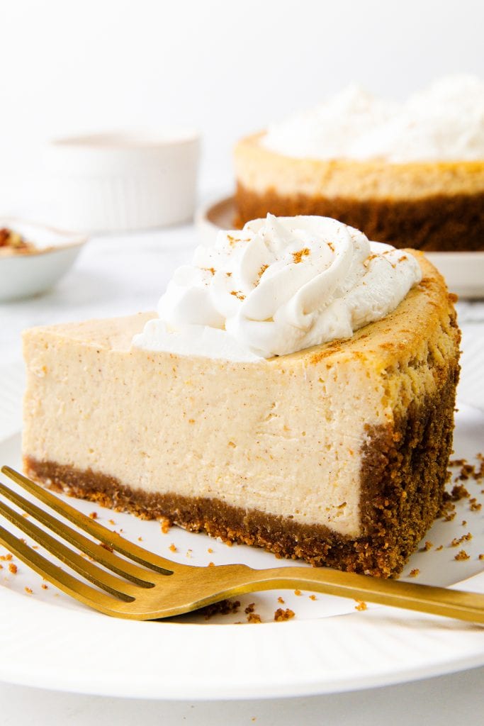 A slice of Pumpkin Spice Cheesecake with a graham cracker crust and a topping of whipped cream sits on a white plate with a gold fork. The rest of the cheesecake is in the background.